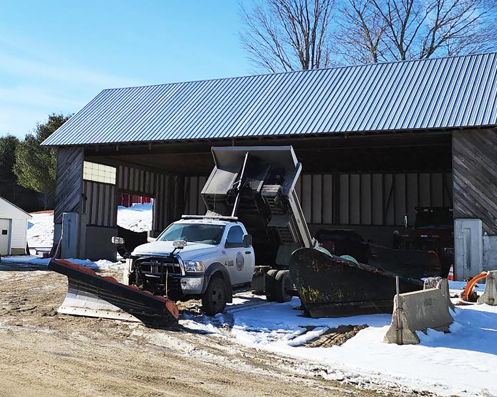 A dump truck from the Town of Unity NH Highway Department is parked near a plow in a storage shelter.