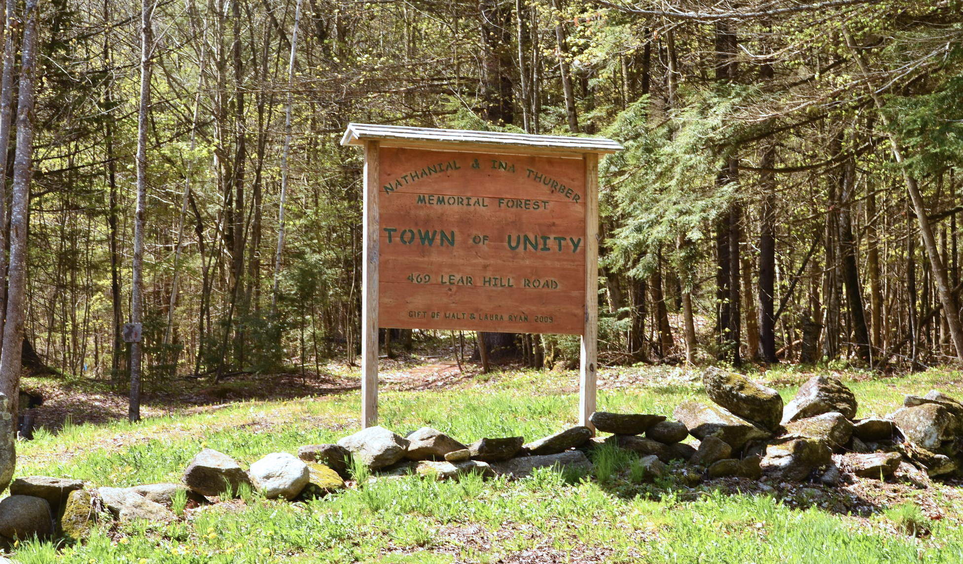 A wooden sign in the Unity NH Thurber Memorial Forest with rocks and trees around it. The sign says Nathanial & Ina Thurber Memorial Forest, Town of Unity.